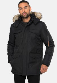 Černá parka s kapucí podšitou kožešinou, několika předními kapsami, zipem a páskovým zapínáním a texturovanou tkaninou.