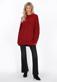 Roter Strickpulli mit gepolstertem Blumenmuster, oversized Passform, Rundhalsausschnitt, kombiniert mit schwarzen Schlagjeans und schwarzen Stiefeletten mit Absatz.
