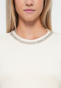 Pull en tricot blanc avec un col rond orné d'une rangée de strass argentés scintillants. Texture douce avec un motif côtelé.