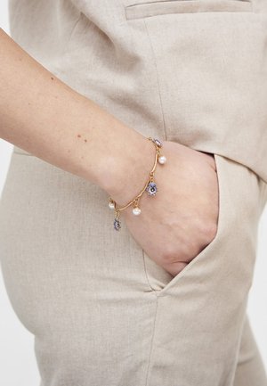 Pols met gouden bedelarmband met paarse bloemen en parels, rustend in de zak van beige linnen broek en bijpassend jasje.