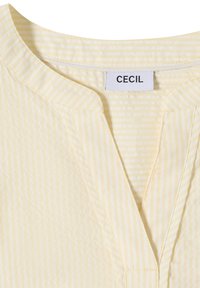 Chemise jaune clair avec de fines rayures verticales, encolure fendue et une étiquette blanche avec l'inscription "CECIL" cousue à l'intérieur du col.