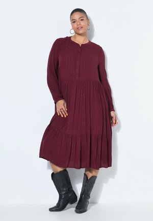 Kobieta ubrana w bordową sukienkę midi z długim rękawem i guzikami oraz czarne kowbojskie buty, stojąca na białym tle.