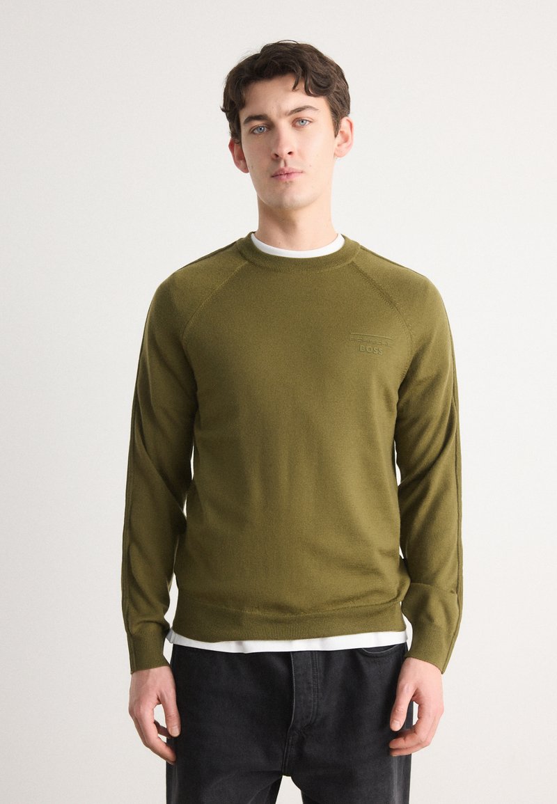 Olijfgroen gebreid sweater met een ronde halslijn, geribbelde manchetten en subtiele logo-detail op de borst, gedragen over een wit shirt.