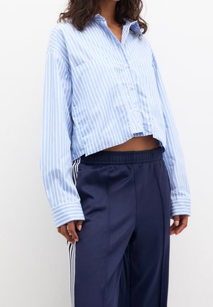 Femme portant une chemise courte rayée bleue et blanche à boutons avec manches longues et un pantalon de survêtement bleu marine avec des bandes blanches sur les côtés.