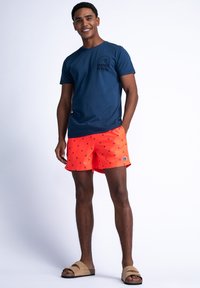 Marineblauwe t-shirt met golfprint, gecombineerd met feloranje zwembroek met kleine blauwe stippen. Bruine slidesandalen maken de look compleet.