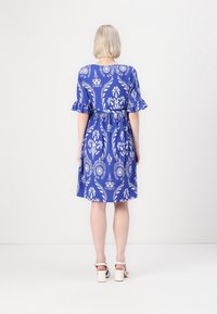 Blå klänning med vit blommönster, korta volangärmar, åtsittande midja och knälång kjol. Bärs med vita klacksandaler.