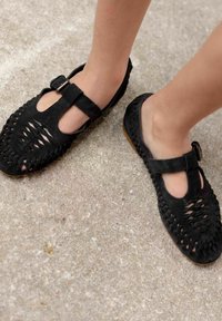 Zapatos negros de ante con tiras en T, detalles de corte, tejido texturizado y una pequeña hebilla plateada. Suela plana, aptos para un uso casual.