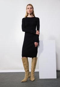 Svart ribbestrikket kjole med lange ermer og gullknappdetaljer ved mansjettene, kombinert med beige semsket semskede knæhøye stiletthæler.