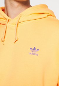 Sudadera amarilla de mezcla de algodón con una textura suave, que presenta un logo de Adidas bordado en púrpura en el pecho y capucha con cordón.