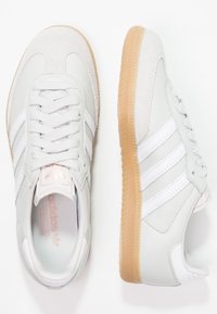 Zapatillas gris claro con una puntera de ante, diseño de tres bandas blancas y suela de goma. Presentan acentos texturizados y cordones planos.