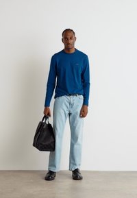 Blå langermet skjorte, lyseblå jeans, svarte sko og en strukturert svart veske. Skjorten har en subtil logo på brystet.