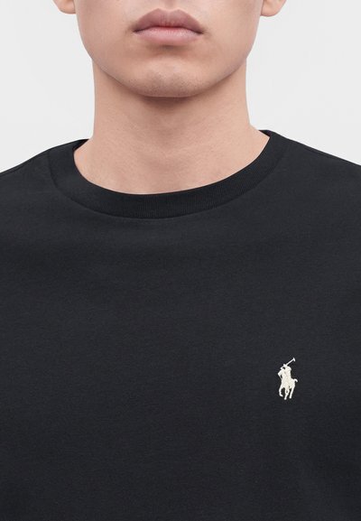 Nærbilde av en persons nedre del av ansiktet og nakken som har på seg en svart genser med rund hals og en liten hvit polo-spillerlogo på brystet.