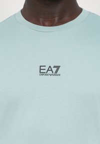Tricou din bumbac de culoare albastru deschis, cu decolteu rotund, având un logo EA7 Emporio Armani de culoare neagră imprimat pe față. Textură netedă.