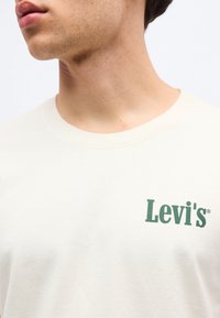 Νεαρός άνδρας που φοράει μια μπλούζα Levi's σε κρεμ αποχρώση με πράσινο λογότυπο στο στήθος, ορατός από το λαιμό μέχρι το μέσο του κορμού σε λευκό φόντο.