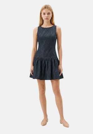 Donna in piedi rivolta in avanti, indossa un abito di jeans scuro senza maniche con gonna svasata e ballerine color nude, contro uno sfondo chiaro e uniforme.