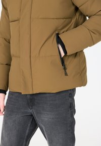 Brun polstret jakke med en lynlåslomme, der har en glat tekstur, sorte detaljer og elastiske manchetter. Båret over mørkegrå jeans.