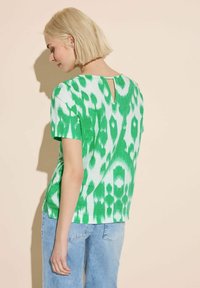 Top corto a maniche corte verde e bianco con motivo, dotato di un dettaglio a chiave sulla schiena, realizzato in un tessuto leggero. Abbinato a jeans blu chiaro.