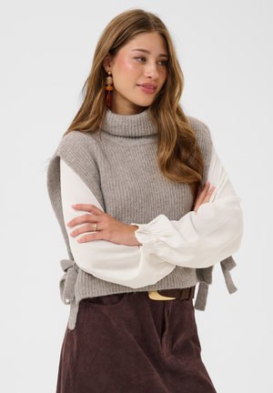 Jeune femme aux longs cheveux portant un col roulé sans manches gris en maille par-dessus un chemisier blanc et un pantalon marron, les bras croisés.