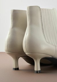 Bottines en cuir beige avec un dos texturé, un talon haut élancé et des détails de couture verticale sur les côtés.