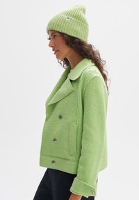 Giacca verde in misto lana con design doppiopetto, ampi rever e grandi bottoni argento. Abbinata a un cuffia a coste coordinata.