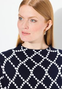 Maglione blu navy con motivo a pois bianchi, scollatura rotonda e tessuto testurizzato. Orecchini a cerchio dorati completano il design.