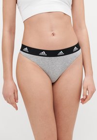 Šedé dámské thong kalhotky s černým elastickým páskem a opakovaným bílým logem Adidas. Hladký povrch, minimalistický design.