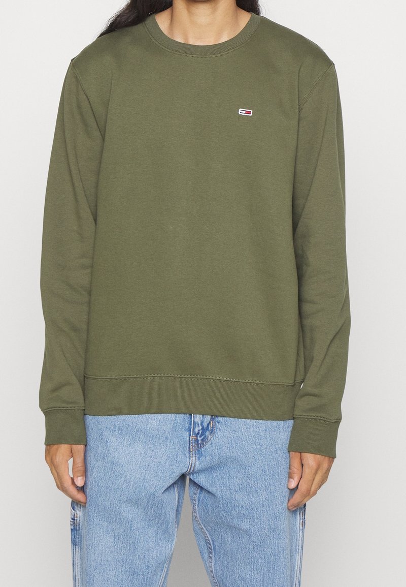 Personne portant un sweat-shirt à manches longues vert olive et un jean bleu clair, debout avec les bras détendus le long du corps.