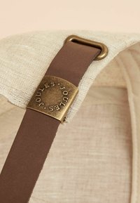 Beigen värinen kangashihna, jossa on ruskeasta nahasta valmistettu yksityiskohta ja kultaisen sävyn metallisolki, johon on kaiverrettu "JOULES" ja jossa on teksturoitu muotoilu.