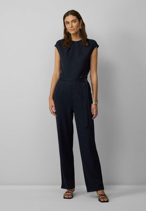 Marineblauer Jumpsuit mit kurzen Ärmeln, weitem Bein und einem gebundenen Taillengürtel. Glatte Stoffoberfläche, kombiniert mit schwarzen Sandalen mit Riemchen.