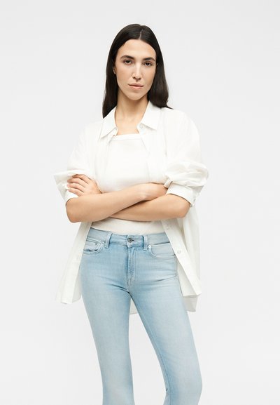 Νεαρή γυναίκα με μακριά σκούρα μαλλιά, φοράει άσπρο πουκάμισο, άσπρο τοπ και ανοιχτό μπλε τζιν, στέκεται με σταυρωμένα τα χέρια πάνω σε απλό φόντο.