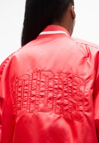 Άτομο με μακριά σκούρα μαλλιά γυρισμένο προς τα πίσω φοράει λαμπερό κόκκινο σατέν μπουφάν με μεγάλο ανάγλυφο κείμενο "DIDAS" στην πλάτη και ριγέ γιακά