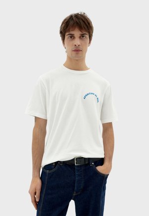 White cotton t-shirt with blue text "APERITIVO AL SOL" on the chest, crew neck, short sleeves, paired with dark blue jeans and black belt.