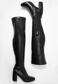 Botas de cuero negras por encima de la rodilla con superficie texturizada, tacón alto y ancho, forma angular y detalle de cierre de cremallera a lo largo del lateral.