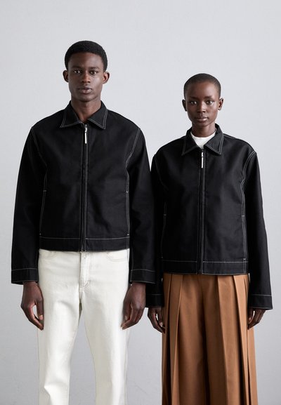 Black cropped jackets with white stitching, front zippers, and pointed collars. One model in cream pants, the other in brown pleated trousers.