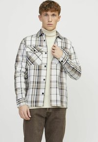 Geruite shirt met een wit, beige en grijs patroon, twee borstzakken, zwarte knopen en lange mouwen, gedragen over een crèmekleurige trui.