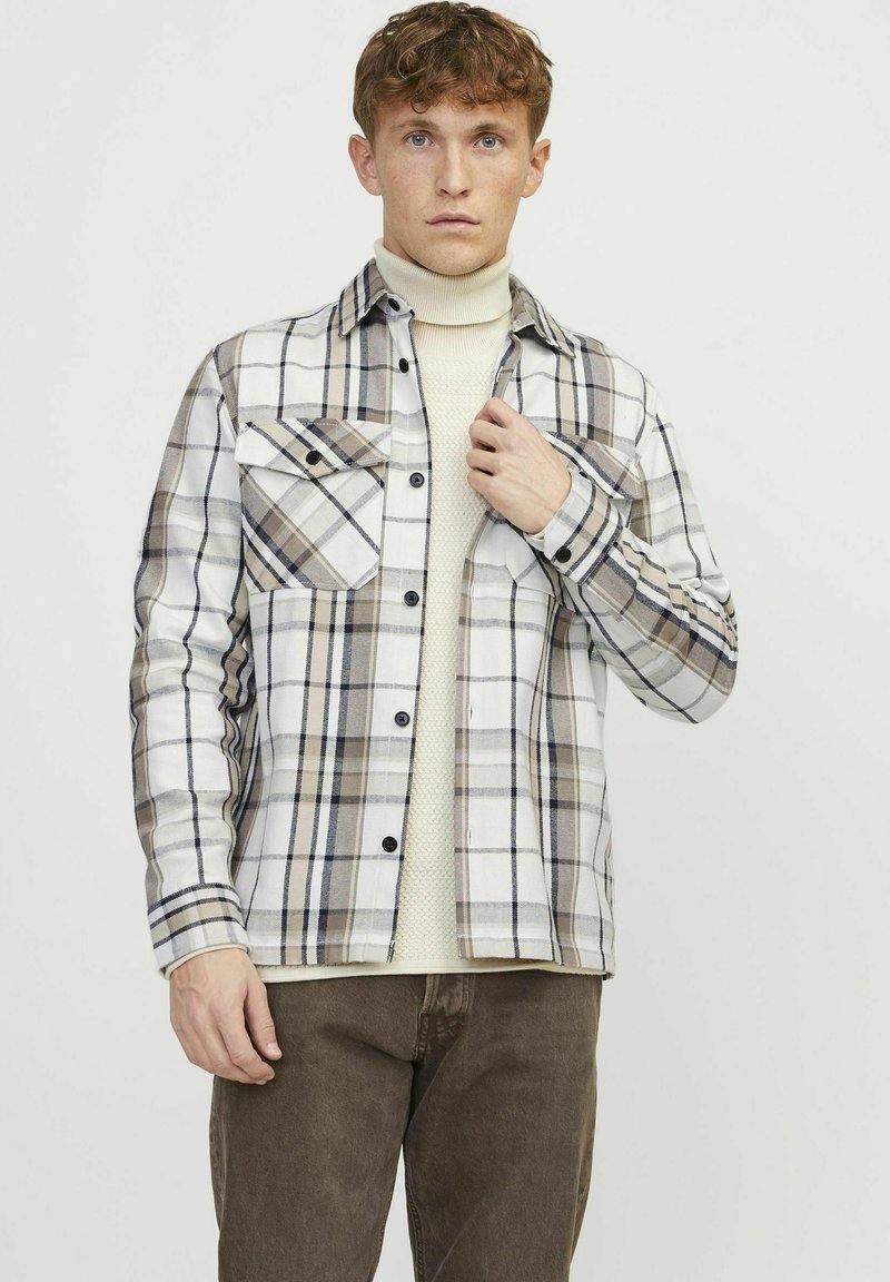 Geruite shirt met een wit, beige en grijs patroon, twee borstzakken, zwarte knopen en lange mouwen, gedragen over een crèmekleurige trui.