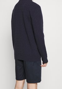 Suéter de punto trenzado en color navy con un cuello alto, patrón texturizado y dobladillo acanalado, combinado con pantalones cortos oscuros. El suéter tiene un corte relajado.