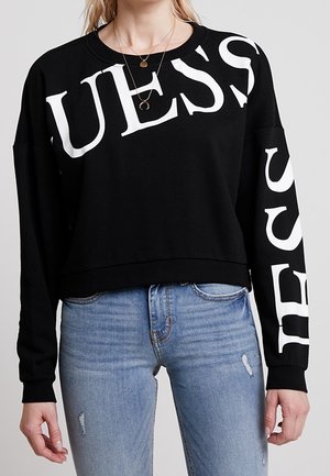 Kvinde iført en sort sweatshirt med stor hvid "GUESS" tekst på brystet og højre ærme, kombineret med lyseblå revnede jeans.