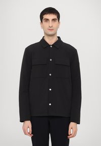Jachetă neagră dintr-un material neted, cu guler, două buzunare frontale și sistem de închidere cu capse. Design simplu și elegant.