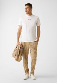 T-shirt bianca con logo "GUESS", abbinata a pantaloni beige con motivo e una giacca beige tenuta in mano, completata da sneakers bianche.