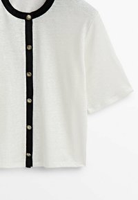 Cardigan blanc à manches courtes avec bordure noire le long de l'encolure et de la patte de boutonnage, avec six boutons dorés décoratifs.