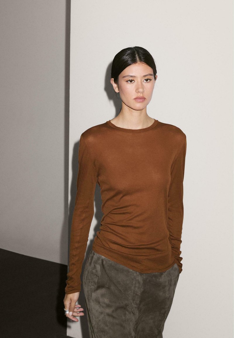 Long-sleeve brown top made of smooth fabric, with a round neckline and fitted sleeves, paired with loose olive green trousers.