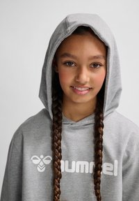 Grauer Kapuzenpullover mit einem großen weißen Logo auf der Brust. Besitzt eine lockere Passform und geflochtenes Haar, das das Gesicht umrahmt.