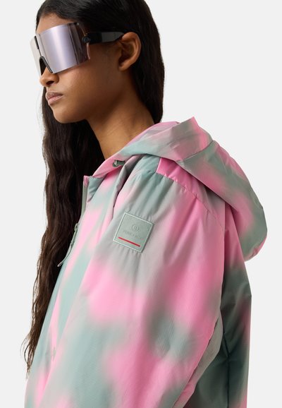 Kvinde med langt mørkt hår iført store reflekterende solbriller og en pink og grøn tie-dye hættetrøje med et Fire + Ice logo-patch.