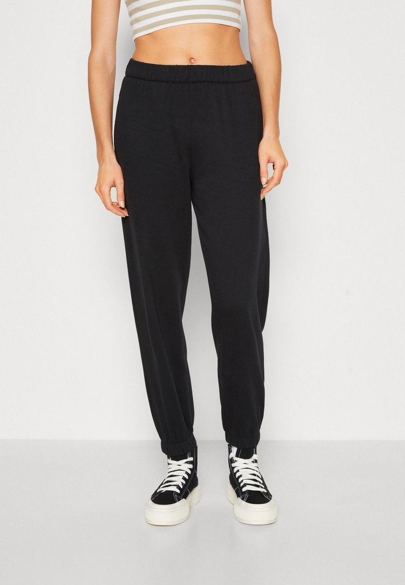 Femme portant un pantalon de jogging noir à taille élastique et des baskets montantes noires avec des lacets blancs, debout devant un fond uni.