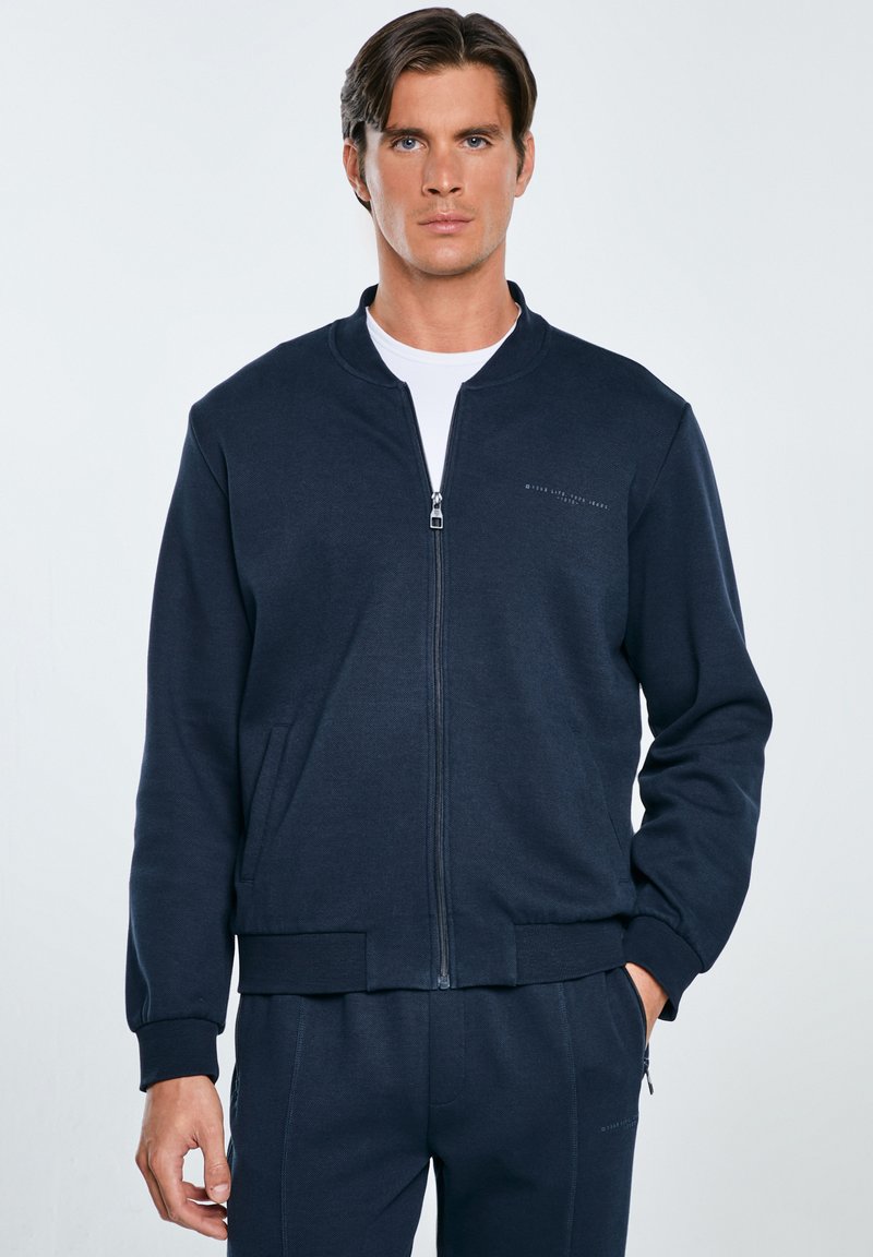 Felpa blu navy con zip, colletto rotondo, tasche laterali, polsini a costine e un logo sottile sul lato sinistro superiore.
