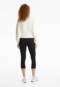Kremasti pulover s dugim rukavima, uparen s crnim capri tajicama. Model nosi svijetle sportske cipele.