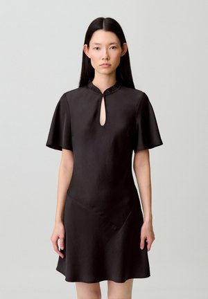 Mujer con cabello largo y oscuro lleva un vestido negro de manga corta con un escote con volantes y abertura en forma de lágrima, de pie contra un fondo claro y liso.