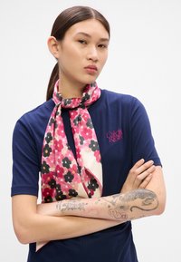 Sciarpa floreale rosa con accenti neri e gialli, realizzata in tessuto morbido, avvolta attorno al collo, abbinata a un top blu navy con un logo.