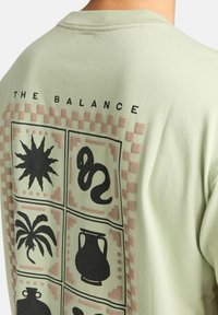 Světle zelené bavlněné tričko se velkým grafickým designem, který obsahuje devět černých ikon s růžovými akcenty, včetně slunce, hada a palmy.